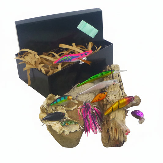 Fishing lure gift set with open black box, multicolor hard lures on rocks and pink feather tail