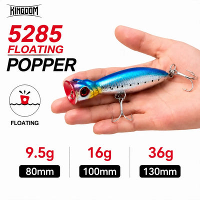 Blue and silver floating popper fishing lure with treble hooks, held in a hand.