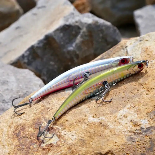 Floating pencil fishing lures on rocks: silver-blue and green-gold striped bodies with treble hooks