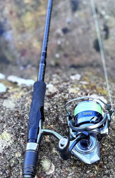 Silver spinning reel with green line spool on rocky shore beside rod